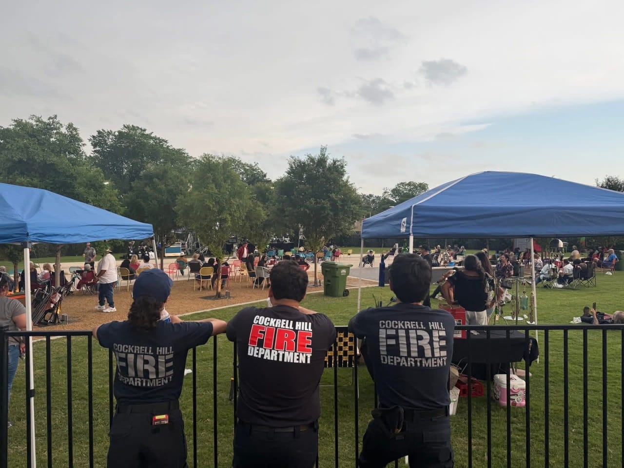 Three CHFD members in department T-shirts watch a community event in a park with canopies and families on the lawn