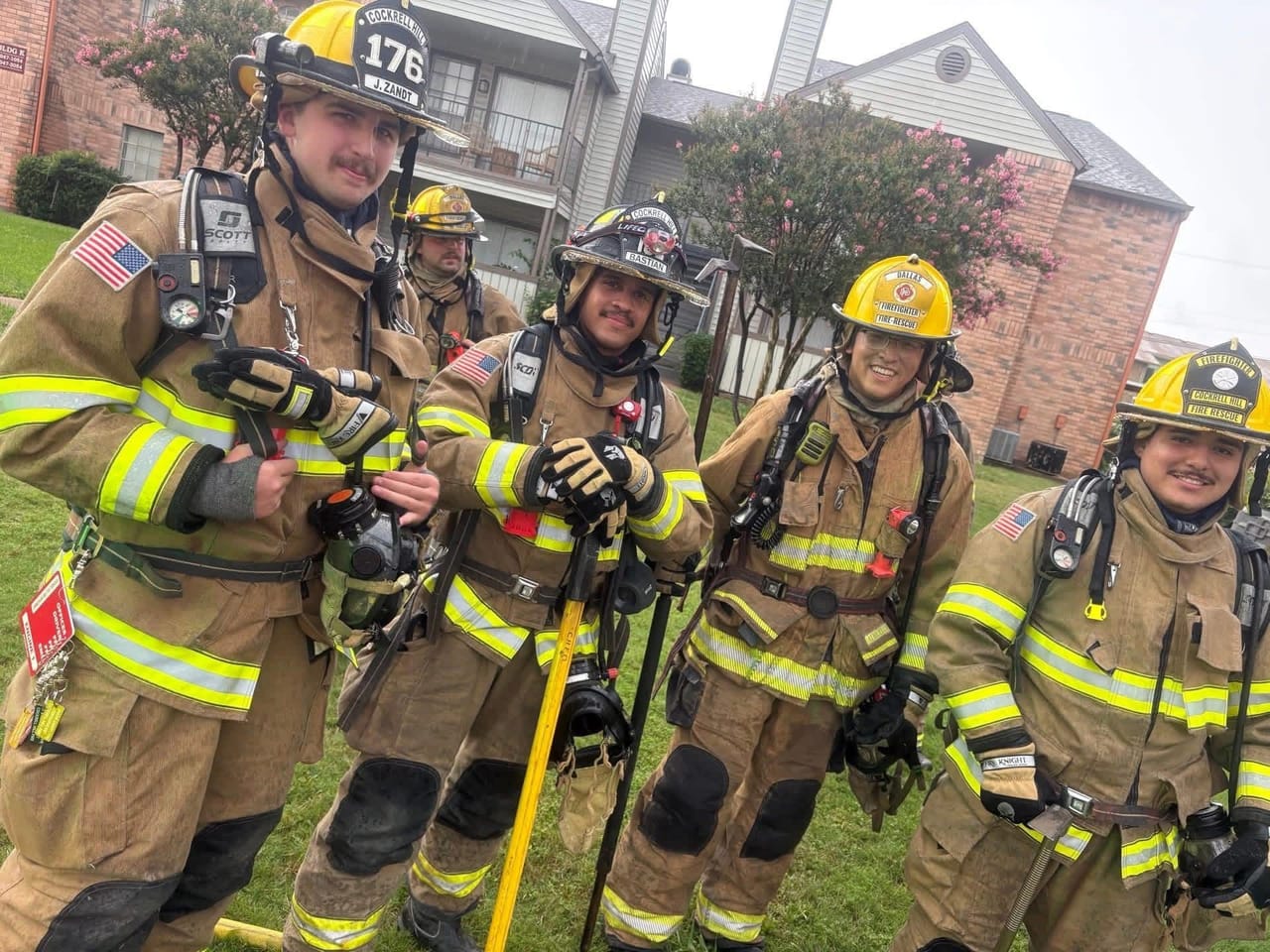Five Cockrell Hill firefighters in full turnout gear and helmets, smiling for the camera outside an apartment complex