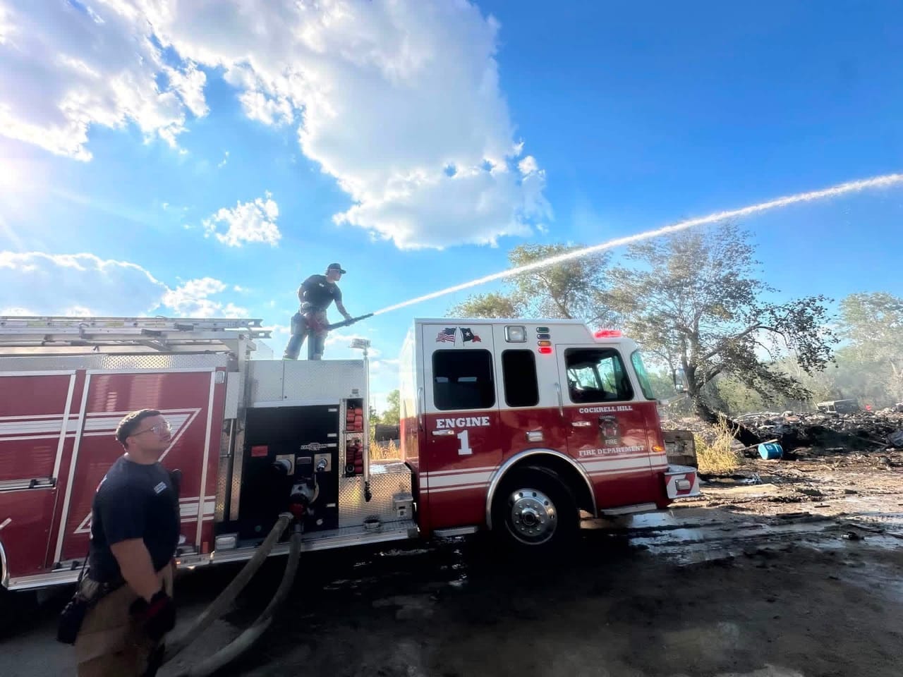 A firefighter on top of Engine 1 directs a stream of water onto a burning debris pile while another works the pump panel