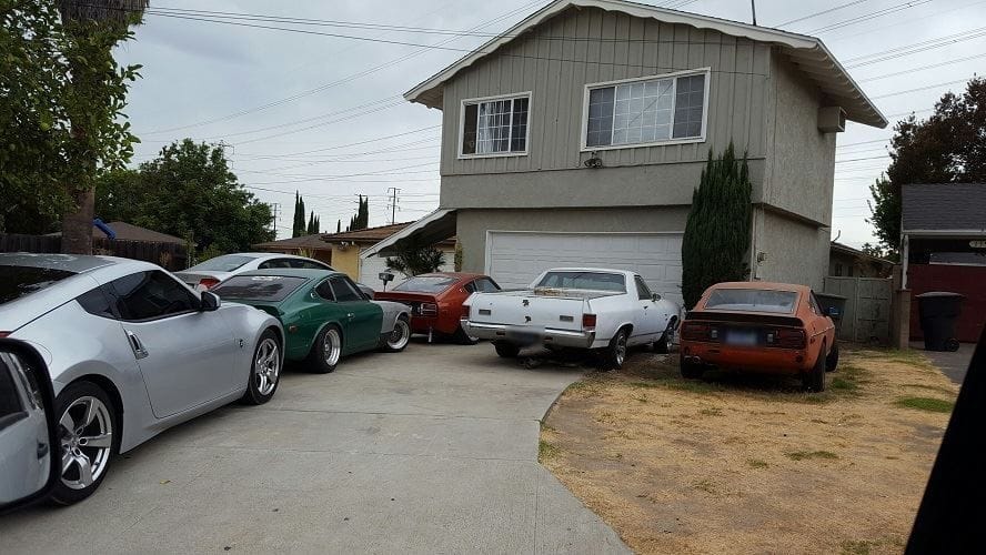 Several cars crowded into a driveway, with an orange car parked on the bare dirt yard in front of a two-story house